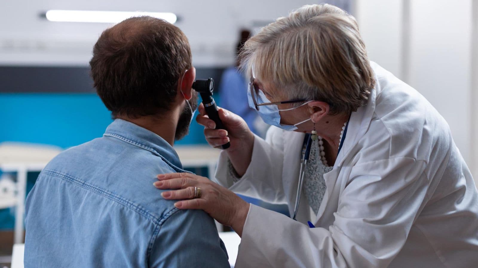 A female doctor wearing a white coat and face mask uses an otoscope to examine the ear of a male patient wearing a light blue denim shirt in clinic.