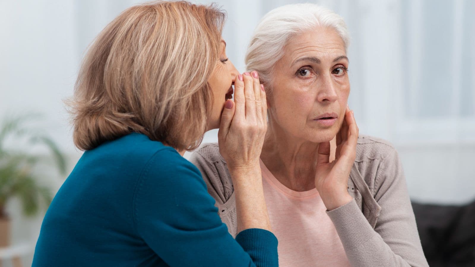 Two women engaged in a conversation, one whispering to the other who looks surprised, in a bright indoor setting.