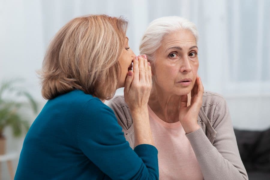 Two women engaged in a conversation, one whispering to the other who looks surprised, in a bright indoor setting.