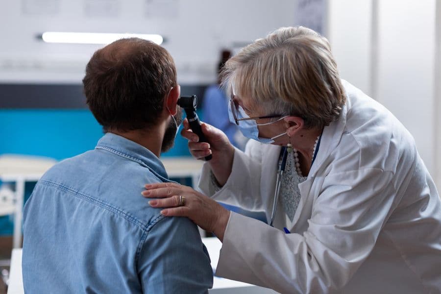A female doctor wearing a white coat and face mask uses an otoscope to examine the ear of a male patient wearing a light blue denim shirt in clinic.